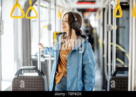 Junge elegante Frau mit Kopfhörer, während in der modernen Straßenbahn bewegt. Glückliche Passagiere genießen Urlaub im öffentlichen Verkehr Stockfoto