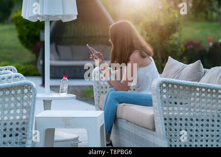 Junge Frau auf einer Terrasse am Abend in einem großen weidensofa Sms eine Nachricht auf ihrem Mobiltelefon mit Hintergrundbeleuchtung durch das Leuchten der Sonne Stockfoto