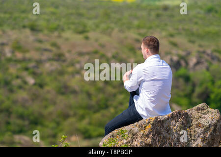 Junge Mann sitzt auf einem hohen Felsen unten mit Blick über die Landschaft, wie er einen erholsamen Tag im Freien in einem gesunden Lebensstil Konzept verbringt Stockfoto