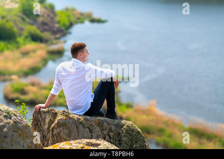 Junge Mann sitzt auf einem erhöhten Felsen auf einem Hügel oder Berg mit Blick auf einen breiten Fluss unten Genießen der Landschaft Stockfoto