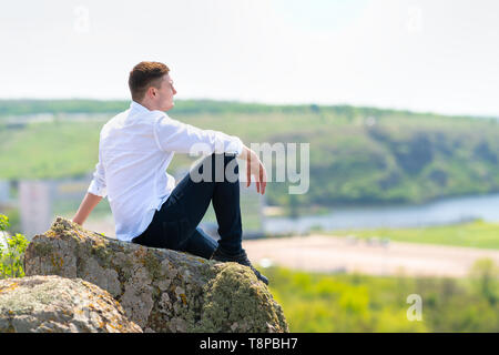 Attraktive intelligente junge Mann sitzt auf einem Felsen mit Blick auf einen Fluss unten in der warmen Sonne in einer Nahaufnahme, Seitenansicht Stockfoto