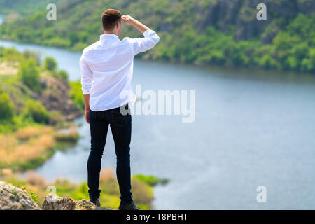 Junge Mann, der auf einem Felsen mit Blick auf einen breiten Fluss mit bewaldeten Ufer unten hob seine Hand in seinen Augen die Sonne zu schützen. Stockfoto