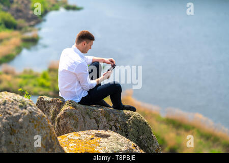 Junger Mann seinem Mobiltelefon für Nachrichten, als er auf einem großen Felsblock mit Blick auf einen breiten Fluss unten in der warmen Frühlingssonne entspannt Stockfoto
