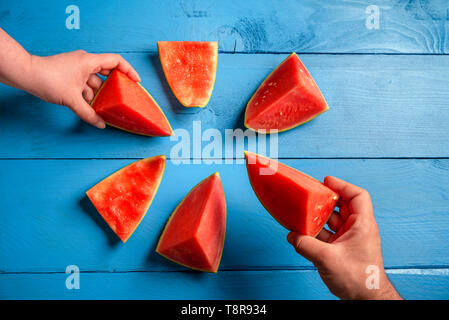 Oben Ansicht von Mann und Frau Hände greifen für Scheiben von roten Wassermelone, über einem blauen Holztisch. Essen Sommer Früchte. Köstliche Sommer behandelt. Stockfoto