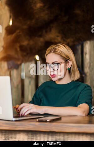 Glücklich Schönheit Friseursalon manager Porträt. Junge blonde Frau Tippen auf Laptop am Schreibtisch am Arbeitsplatz. Sie tragen grüne Sweatshirt und schwarzer Brille, sm Stockfoto