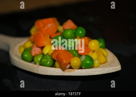 Stücke von Karotten, mais und Erbsen auf hölzernen Löffel mit dunklen Hintergrund Stockfoto