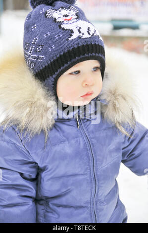 Baby im Winter Overalls in ein Tuch eingewickelt Spaziergänge auf eine verschneite Straße und formt aus Schnee. Stockfoto