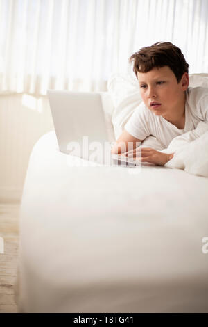 Pre-heranwachsenden Jungen spielt mit einem Computer mit einem Laptop spielt, während im Bett in ein weißes Schlafzimmer liegen. Stockfoto