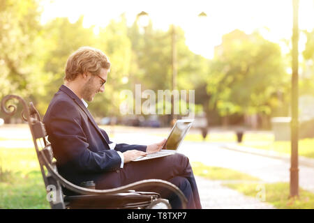 Hübscher junger Geschäftsmann mit Laptop auf der Bank im Park Stockfoto