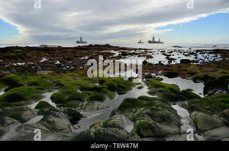 An der felsigen Küste bei Ebbe mit kleinen Pfützen im Vordergrund und Ölplattformen im Hintergrund Stockfoto