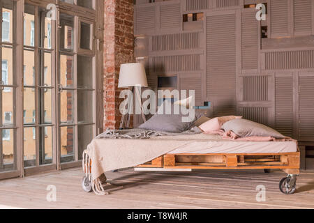 Loft Apartment Interieur. Minimalistische Einrichtung mit einfachen Bett aus Holz, Lampe, großes Fenster. Stilvolle Schlafzimmer im Loft Stil in Pastelltönen gestaltet. Platz kopieren Stockfoto