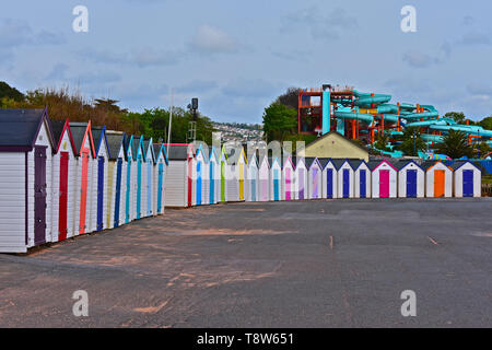 Eine bunte Reihe von bunten Badekabinen an der Strandpromenade mit Blick auf das Meer bei Goodrington Sands. Blaue Folien auf den nahe gelegenen Wasserpark hinter sich. Stockfoto