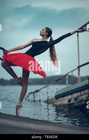 Ballerina tanzt an der Küste des Flusses in einem schwarzen und roten Tutu gegen den Hintergrund der alten Boot. Stockfoto