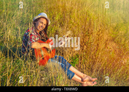 Schöne, glückliche Frau im Sommer in den Strahlen der untergehenden Sonne mit einer Gitarre. Konzept der kreativen Freiheit, Spaß und Ferien Stockfoto