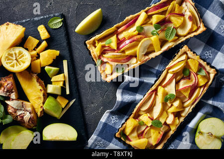 Leckere Ananas und Apfel süße Torte in rechteckigen Tart Pfannen auf einer konkreten Tabelle mit Küchentuch und Zutaten, horizontale Ansicht von oben, Stockfoto