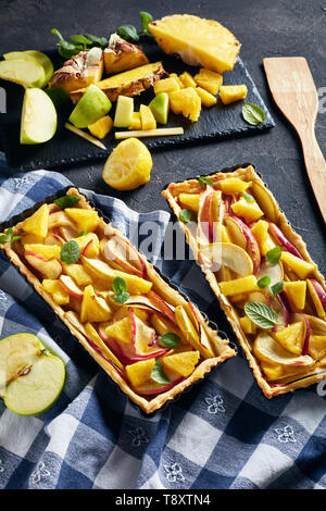 Leckere Ananas und Apfel süße Torte in rechteckigen Tart Pfannen auf einer konkreten Tabelle mit Küchentuch und Zutaten, vertikale Ansicht von oben Stockfoto