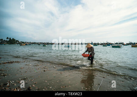 Vietnamesische Frau in einem kegelförmigen Hut wäscht Schalentiere in einem Korb im Meer. MUI ne. Vietnam. 27.01.2019 Stockfoto