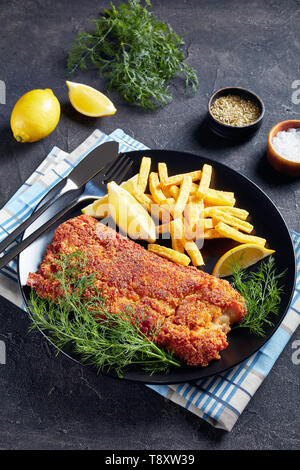 Close-up Paniertes Seehechtfilet mit Chips, frischer Dill und Zitronenscheiben auf schwarzem Teller serviert auf einer konkreten Tabelle, vertikale Ansicht von oben Stockfoto