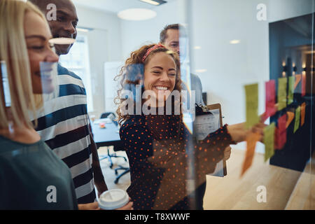 Lächelnden jungen Geschäftsfrau lesen Haftnotizen auf eine Glaswand beim Brainstorming mit Kollegen im Büro Stockfoto