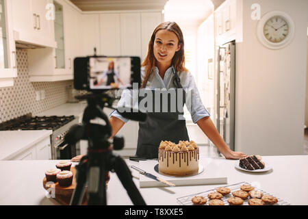 Junge Frau vlogger Backen und Aufnahme von Video für Lebensmittel. Weibliche Konditor vlogging mit Ihrem Mobiltelefon auf einem Stativ in der Küche montiert. Stockfoto