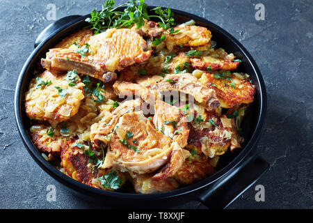 Lecker gebräunt Schweinekoteletts auf den Knochen- und Hash Brown Eintopf in einem schwarzen Keramik pan auf einer konkreten Tabelle, horizontale Ansicht von oben, close-up Stockfoto