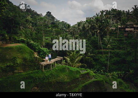 Paar steht auf hölzerne Brücke in der Nähe von Reisterrassen auf Bali, Indonesien. Halten sich an den Händen. Romantische Stimmung. Urlaub in den Tropen. Luftaufnahme. Auf Hintergrund Stockfoto