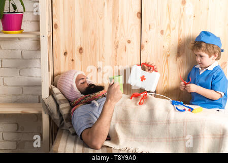 Kind und Vater spielen Krankenhaus. Patienten mit Grippe im Bett lag, unter Wolldecken. Arzt besuchen Kranke bärtiger Mann zu Hause, Lernspiel Stockfoto