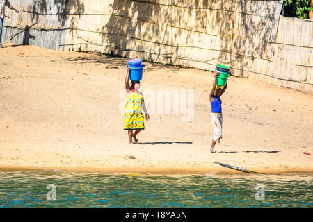 Zwei junge MALAWISCHE Frauen mit Eimer auf dem Kopf kommen zum Lake Malawi um Wasser zu zeichnen Stockfoto