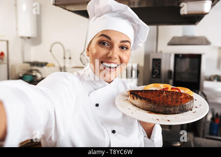 Porträt der lächelnde Frau Koch tragen weiße Uniform unter selfie Foto halten Sie die Platte mit dem Essen in der Küche im Restaurant Stockfoto