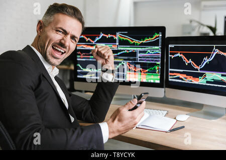 Portrait der männlichen Mitarbeiter 30 s tragen mit Handy während der Arbeit mit digitalen Grafiken am Computer in Bürosoftware Stockfoto