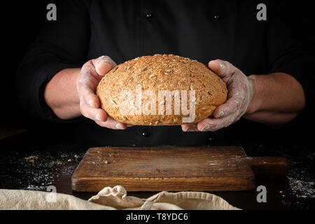 Koch in schwarzen Uniform hält ganze runde Brot mit Sonnenblumenkernen über Holz- Tabelle Stockfoto