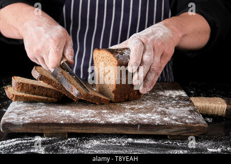 Koch in schwarzen Uniform hält ein Küchenmesser in der Hand und schneidet Stücke Brot aus einer gebackenen braun Roggenmehl Brot auf einem Holzbrett Stockfoto