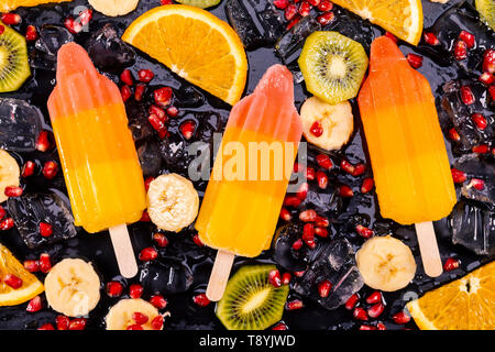 Obst Eis auf Stick mit Scheiben Früchte auf schwarzem Schiefer. Fokus auf Eis am Stiel. Stockfoto