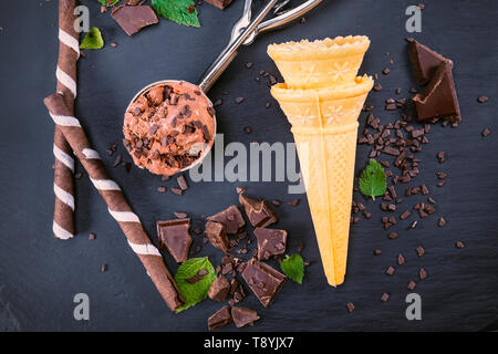 Schokoladeneis in Schaufel mit wafer Sticks, Kegel und Schokolade auf schwarzem Schiefer. Fokus auf Eis in Schaufel und auf Wafer Kegel. Stockfoto