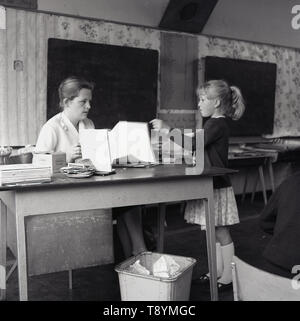 1960er, historisch, Grundschulmädchen, die mit ihrem Lehrer spricht, der an ihrem Schreibtisch im Klassenzimmer sitzt, England, Großbritannien. Stockfoto