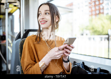 Junge und glückliche Frau mit Smartphone, während in der Nähe der Fenster in den öffentlichen Verkehrsmitteln sitzen während der Fahrt Stockfoto