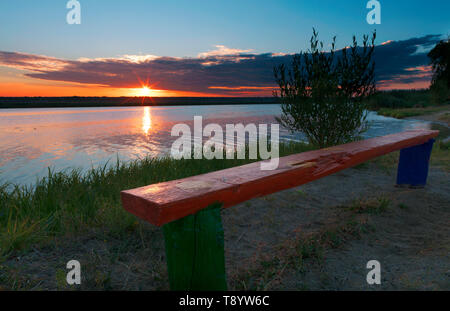 Die hellen aufgehende Sonne beleuchtet die Küsten Gras, eine alte Holzbank und Büsche wachsen auf dem Fluss Stockfoto