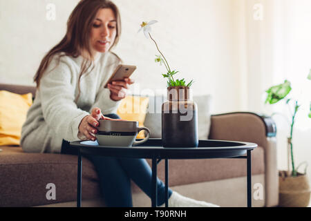 Junge Frau, die sich in Wohnzimmer mit Smartphone und Kaffee zu trinken. Home Inneneinrichtung Stockfoto