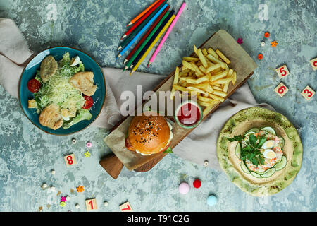 Auf einem grauen strukturierten Hintergrund ist ein Blauer Schild mit tseat Salat, ein Holzbrett mit einem Burger und Pommes frites, ein gelbes Schild mit Krabben Salat. Schöne Gerichte. Kindermenü im Restaurant. Ansicht von oben Stockfoto