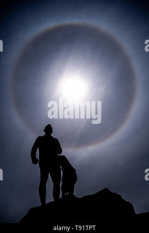 Aberystwyth Wales UK, Donnerstag, den 16. Mai 2019 Deutschland Wetter: Ein Mann und sein Hund arr so perfekt kreisförmigen Sonne halo silhouetted, gebildet, wie die Strahlen der Sonne durch Eiskristalle in der oberen Atmosphäre, füllt sich der Himmel über Aberystwyth. Die Halos, immer auf 22º von der Sonne, häufig darauf hin, dass Regen wird innerhalb der nächsten 24 Stunden fallen, da die cirrostratus Wolken, die sie verursachen, kann ein sich näherndes Frontschutzsystem bedeuten. Das Wetter wird eingestellt über Nacht aus dem feinen warmen Bedingungen zum kühler und feuchter Tage, eher typisch für Mitte Mai. Photo credit Keith Morris/Alamy Leben Nachrichten zu ändern. Stockfoto