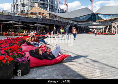 Menschen auf roten Kissen mit einem Bett von leuchtend roten Blüten im kürzlich eröffneten öffentlichen Raum an Kohle Tropfen Hof, King's Cross, London, UK, 2019 Stockfoto