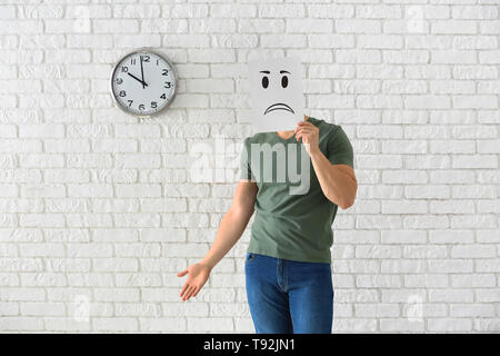 Junger Mann verstecken Gesicht hinter Blatt Papier mit gezogenen Emoticon gegen weiße Mauer mit Uhr Stockfoto