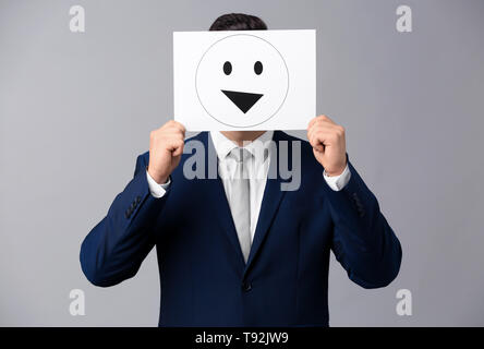 Geschäftsmann ausblenden Gesicht hinter Blatt Papier mit gezogenen Emoticon auf grauem Hintergrund Stockfoto