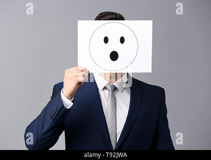 Geschäftsmann ausblenden Gesicht hinter Blatt Papier mit gezogenen Emoticon auf grauem Hintergrund Stockfoto