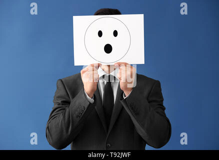 Geschäftsmann ausblenden Gesicht hinter Blatt Papier mit gezogenen Emoticon auf farbigen Hintergrund Stockfoto