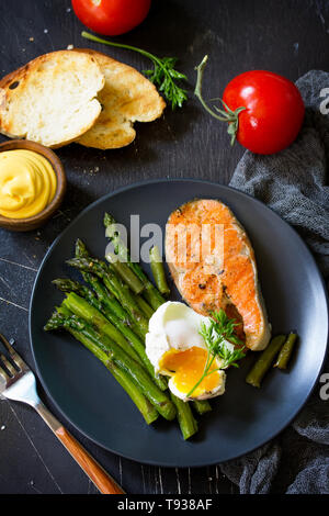 Lachs Fisch Steak vom Grill mit Spargel, pochierte Eier auf schwarzem Hintergrund. Gesundes Essen. Stockfoto