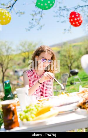 Jugendlich im Alter von Mädchen in rot kariertem Hemd sitzt von Tabelle auf Geburtstag Gartenparty Essen mit Kunststoff Gabel Stockfoto