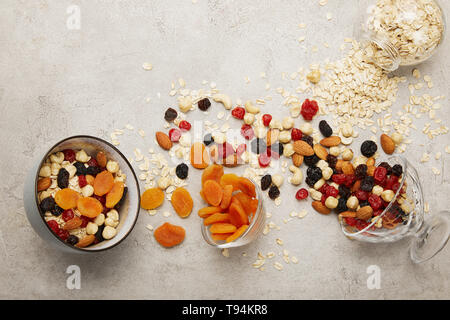 Blick von oben auf die Schüsseln mit Müsli, getrocknete Aprikosen und Beeren, Nüsse auf strukturierte graue Oberfläche mit unordentlich verstreut Zutaten Stockfoto