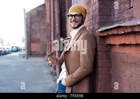 Hübscher junger Mann in Freizeitkleidung holding Einweg Becher und lächelnd während zu Fuß durch die Stadt. Stockfoto