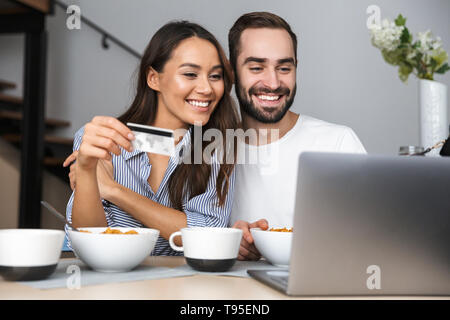 Happy multiethnischen Paar Frühstück in der Küche, am Laptop suchen, mit Kreditkarte Stockfoto